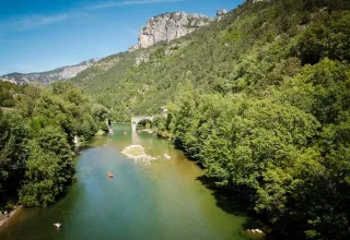 Fluss mit bewaldeten Hängen und einer Steinbrücke nahe Lanuéjols in Okzitanien, Frankreich, bei Sonnenschein.