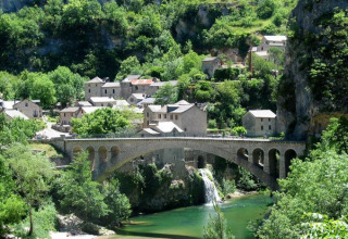 Steinbrücke über einen Fluss mit Wasserfall, umgeben von alten Häusern bei Lanuéjols in Okzitanien, Frankreich.
