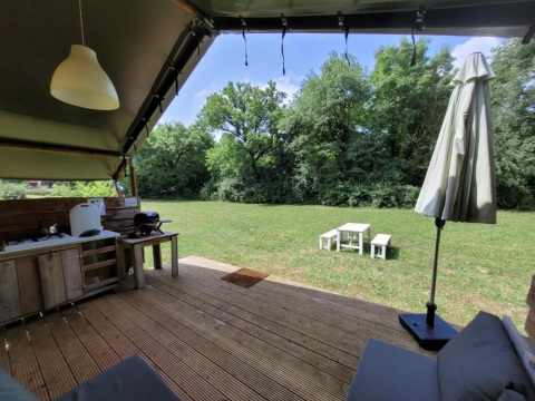 Vista da una terrazza in legno con cucina esterna su prato verde e tavolo da picnic in campeggio soleggiato.