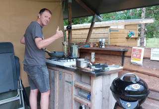 Man cooking at an outdoor kitchen in a safari tent camp, giving a thumbs up towards the camera.