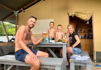 Gezin geniet buiten bij een safaritent op Camping Mas du Midi in Frankrijk, zittend aan tafel met eten en drinken.