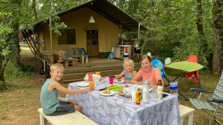 Famille prenant un repas devant une tente safari au camping Mas du Midi en France, entourée de forêt.