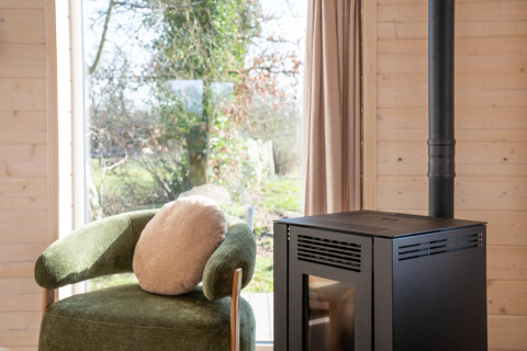 Interieur van Slowlodge bij Zomerlicht in Nederland met groene stoel, kussen en zwarte haard bij het raam.