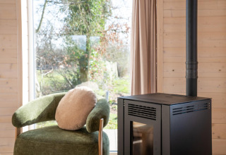 Interieur van Slowlodge bij Zomerlicht in Nederland met groene stoel, kussen en zwarte haard bij het raam.