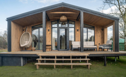 Slowlodge houten lodge met zithoek op de veranda, hangstoel en grote glazen deuren in een groene omgeving.