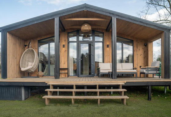 Slowlodge houten lodge met zithoek op de veranda, hangstoel en grote glazen deuren in een groene omgeving.