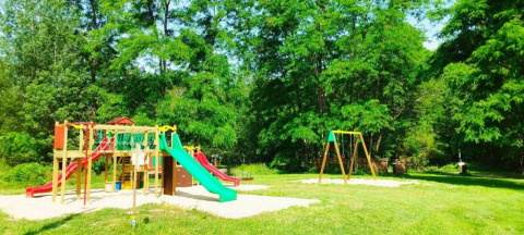 Parco giochi con altalene e scivoli tra alberi verdi al campeggio La Prade, Occitania, Francia.