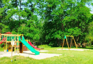 Parco giochi con altalene e scivoli tra alberi verdi al campeggio La Prade, Occitania, Francia.