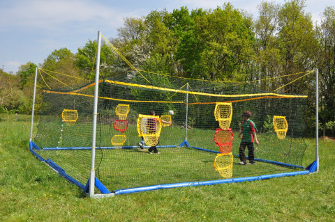 Due bambini giocano in un'area sportiva recintata da reti con bersagli colorati, circondati da erba e alberi.