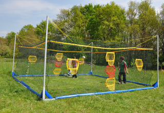 Due bambini giocano in un'area sportiva recintata da reti con bersagli colorati, circondati da erba e alberi.