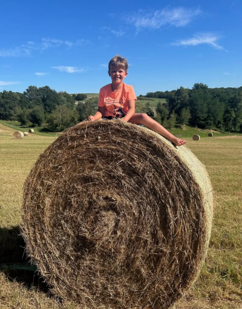 Boy sitting on a large hay bale in Domaine d'Essendieras, Nouvelle-Aquitaine, France, with fields and bales behind.