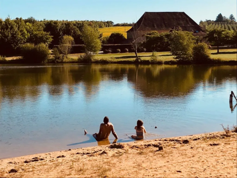 Twee personen zitten aan de oever van het meer bij Domaine d' Essendieras met huis en natuur op de achtergrond.