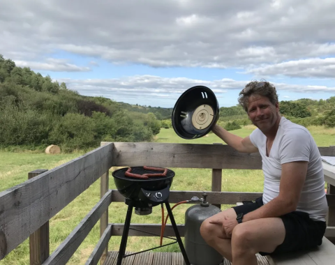 Man grilling sausages on a terrace with valley views at Safaritent met badkamer, Domaine d'Essendieras, France.