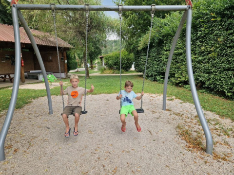 Due bambini seduti sulle altalene in un campeggio a Bourgogne-Franche-Comté, Francia, circondati dal verde.