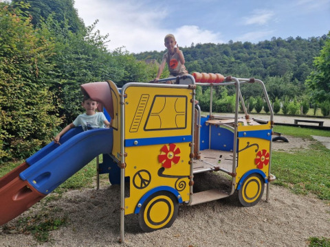 Due bambini giocano su un colorato parco giochi a forma di autobus con scivolo, circondati dal verde.