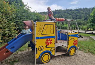Due bambini giocano su un colorato parco giochi a forma di autobus con scivolo, circondati dal verde.