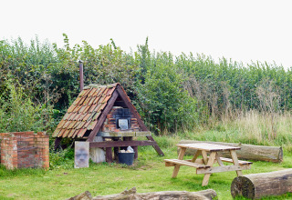 Picknickbereich im Freien mit überdachtem Ofen, Holztisch und Bänken auf Wiese bei BoerenBed College Farm.