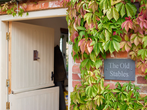 Indgang til The Old Stables med åbnet stalddør og mur dækket af grønne og røde klatreplanter i England.