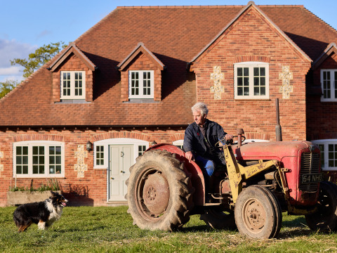 Oudere man op een rode tractor voor een bakstenen huis met hond ernaast op BoerenBed Foxglove farm.
