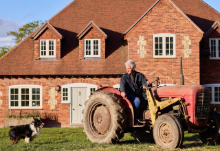 Oudere man op een rode tractor voor een bakstenen huis met hond ernaast op BoerenBed Foxglove farm.