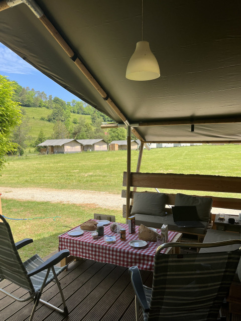 Safari tent porch with outdoor seating, table with red checkered cloth, facing grassy field and tents.