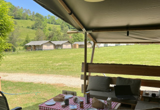 Véranda de tente safari avec sièges extérieurs, table à nappe à carreaux rouges, face à un pré et des tentes.
