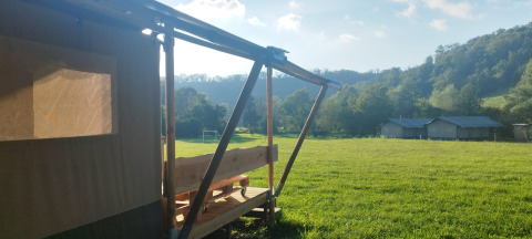 View from a safari tent porch overlooking a green meadow with hills and trees in the sunny distance.