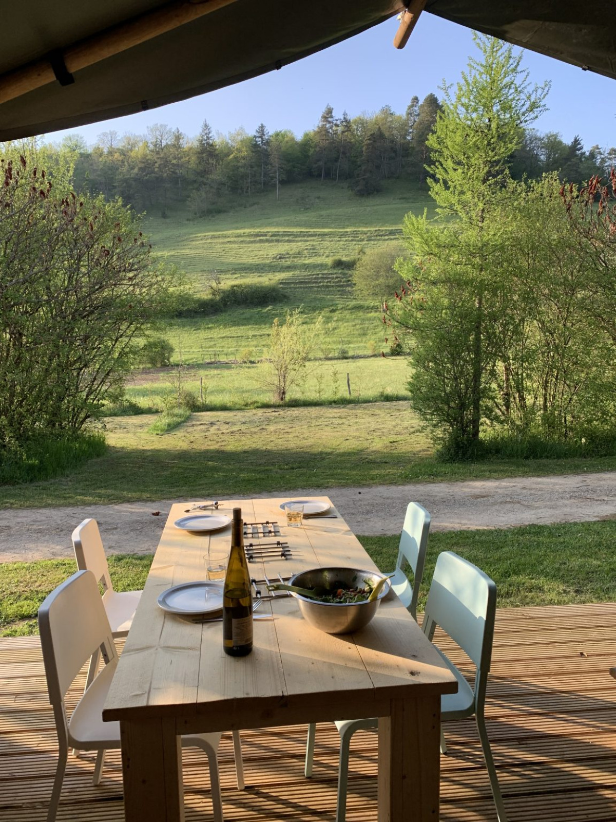 Houten buitentafel met vier stoelen, wijnfles, slakom en uitzicht op groene velden en bomen.