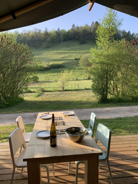 Table de salle à manger en bois à l'extérieur, quatre chaises, bouteille de vin, salade et vue verdoyante.