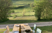 Outdoor wooden dining table with four chairs, wine bottle, salad bowl, and a view of fields and trees.