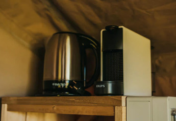 Electric kettle and KRUPS coffee machine on a wooden shelf, pictured inside a safari tent with soft light.