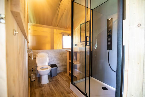 Safari tent bathroom with a toilet, modern glass shower, wooden shelving, and bright natural lighting.