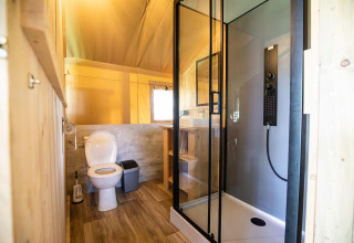 Safari tent bathroom with a toilet, modern glass shower, wooden shelving, and bright natural lighting.