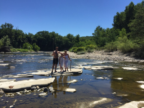 Una famiglia in piedi sulle rocce in un fiume poco profondo al Campeggio La Digue, Auvergne-Rhône-Alpes, alberi verdi.