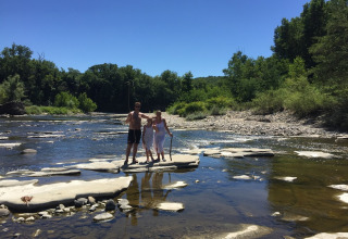 Una famiglia in piedi sulle rocce in un fiume poco profondo al Campeggio La Digue, Auvergne-Rhône-Alpes, alberi verdi.