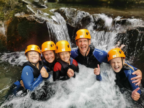 Famiglia con caschi e mute che si diverte sotto una cascata al campeggio La Digue, Auvergne-Rhône-Alpes, Francia.