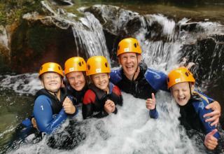 Famiglia con caschi e mute che si diverte sotto una cascata al campeggio La Digue, Auvergne-Rhône-Alpes, Francia.