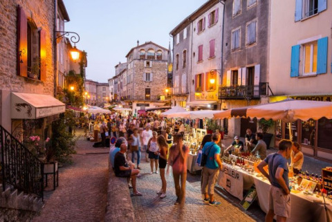 Vivace mercato serale nelle strade di Ruoms, Francia, con bancarelle illuminate e folla passeggiando.