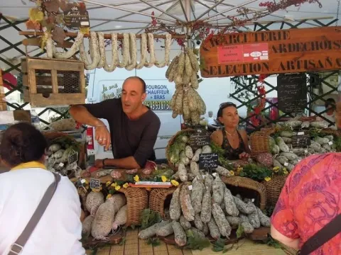 Scena di mercato vicino a Ruoms, Francia, con venditori locali e una vasta esposizione di salumi artigianali.