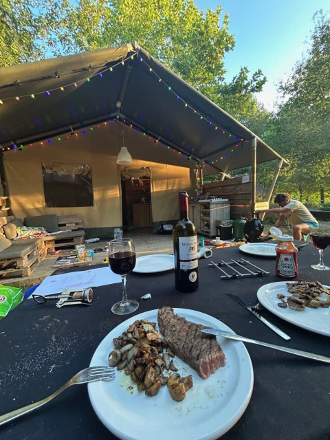 Abendessen vor einem Safarizelt mit gebratenem Fleisch, Wein und bunten Lichterketten in der Natur.
