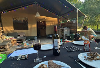 Cena junto a una tienda de safari con carne asada, papas, vino tinto y luces coloridas en la naturaleza.