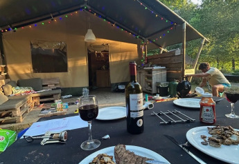 Abendessen vor einem Safarizelt mit gebratenem Fleisch, Wein und bunten Lichterketten in der Natur.
