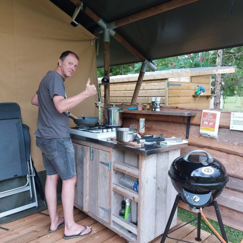 Mann kocht in einer Outdoor-Küche eines Safarizelts, Daumen hoch, entspannte Campingatmosphäre.