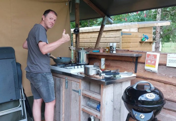Mann kocht in einer Outdoor-Küche eines Safarizelts, Daumen hoch, entspannte Campingatmosphäre.