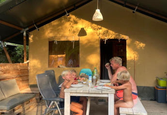 Familie sidder udenfor og spiser ved et bord foran en Safaritent om sommeren i skoven.