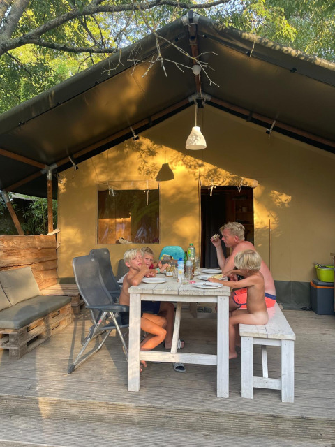 Familia comiendo junta al aire libre frente a una tienda de safari rodeada de naturaleza y sol.