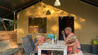 Famiglia che mangia insieme all’aperto davanti a una tenda safari immersa nella natura d’estate.
