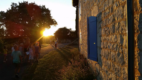 Bambini e adulti passeggiano al tramonto accanto a un edificio in pietra con persiane blu in Occitania, Francia.
