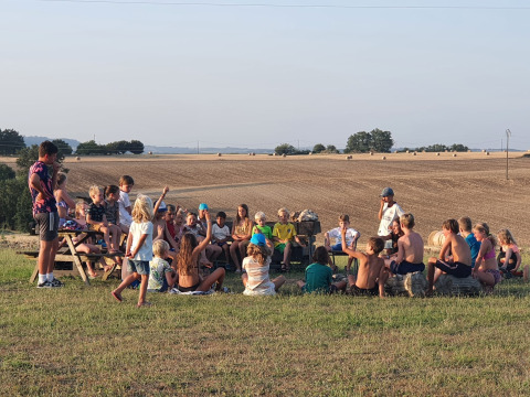 Bambini seduti in cerchio sull’erba al Camp Redon, Occitania, Francia, con campi sullo sfondo panoramico.