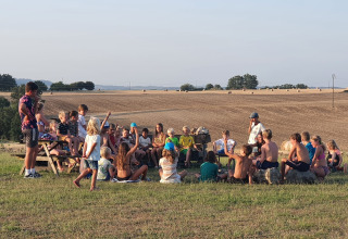 Bambini seduti in cerchio sull’erba al Camp Redon, Occitania, Francia, con campi sullo sfondo panoramico.